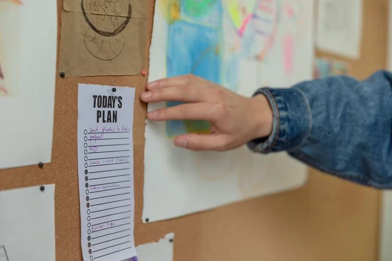 A parent pinning a daily plan next to a child's artwork while planning for school days off